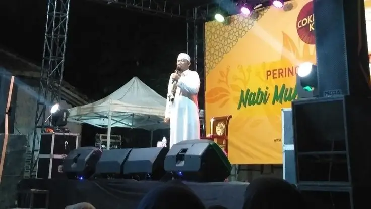 Agama di Panggung Publik  Bitchology Seorang ulama berdiri di atas panggung dengan sorotan lampu menggambarkan bagaimana agama tampil di ruang publik modern sebagai tontonan sosial