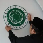 Seorang Muslim sedang menempelkan plang bertuliskan MUI di dinding menggambarkan simbol lembaga keagamaan yang berjarak dari semangat keterbukaan dan dialog